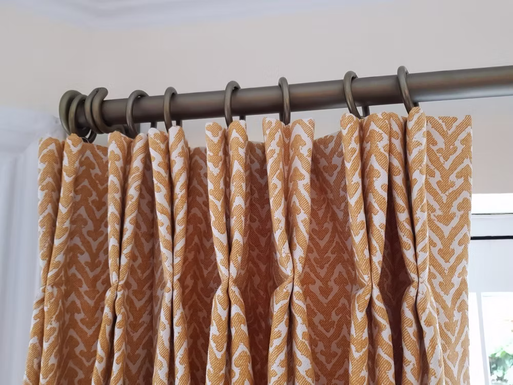An ochre and white geometric pinch-pleated curtain hanging from an antique brass pole with matching rings, beside a white-framed window