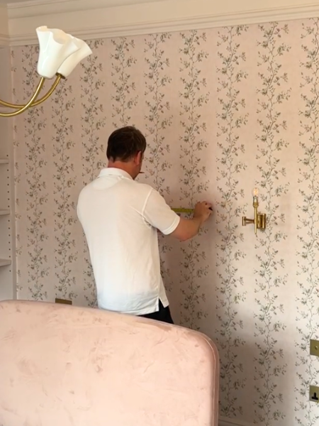 A Chatelaine installer measuring curtain hardware on a floral wallpapered bedroom wall