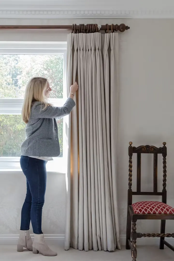 A Chatelaine maker dressing full-height pleated curtains on a wooden pole in a client's bedroom