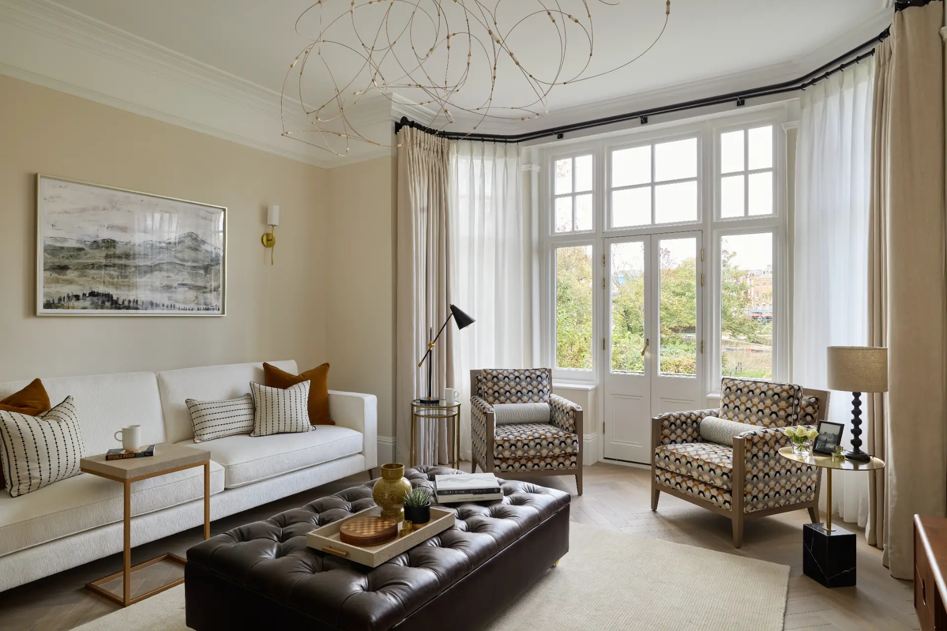 A bright period living room with full-height curtains and voiles, a cream sofa and patterned armchairs