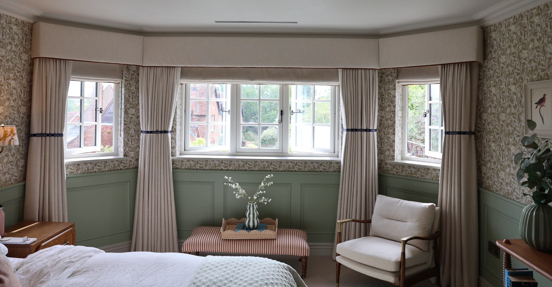 A bay window dressed with a plain oatmeal upholstered pelmet with piped trim, gathered curtains on branded ribbon tie-backs and matching roman blinds, against botanical wallpaper and sage paneling